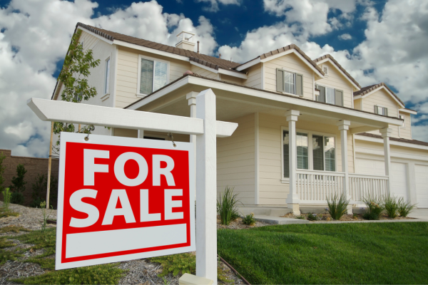 Suburban two-story beige house with front porch and garage, featuring a red “For Sale” sign in the yard. Suburban two-story beige house with front porch and garage, featuring a red “For Sale” sign in the yard.