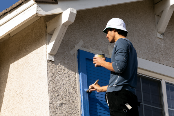 Man painting blue shutters on a house exterior with a brush and paint can Man painting blue shutters on a house exterior with a brush and paint can