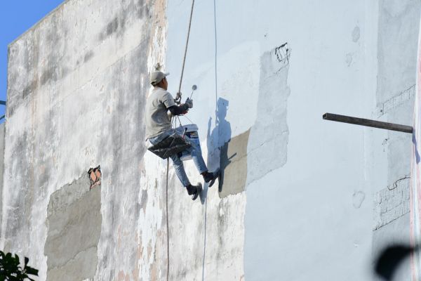 A painter suspended by a harness works on a tall, weathered exterior wall, applying fresh paint while holding a bucket and tools. A painter suspended by a harness works on a tall, weathered exterior wall, applying fresh paint while holding a bucket and tools.